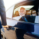 Business traveler exiting a luxury vehicle with chauffeur assistance for corporate car service to the airport.
